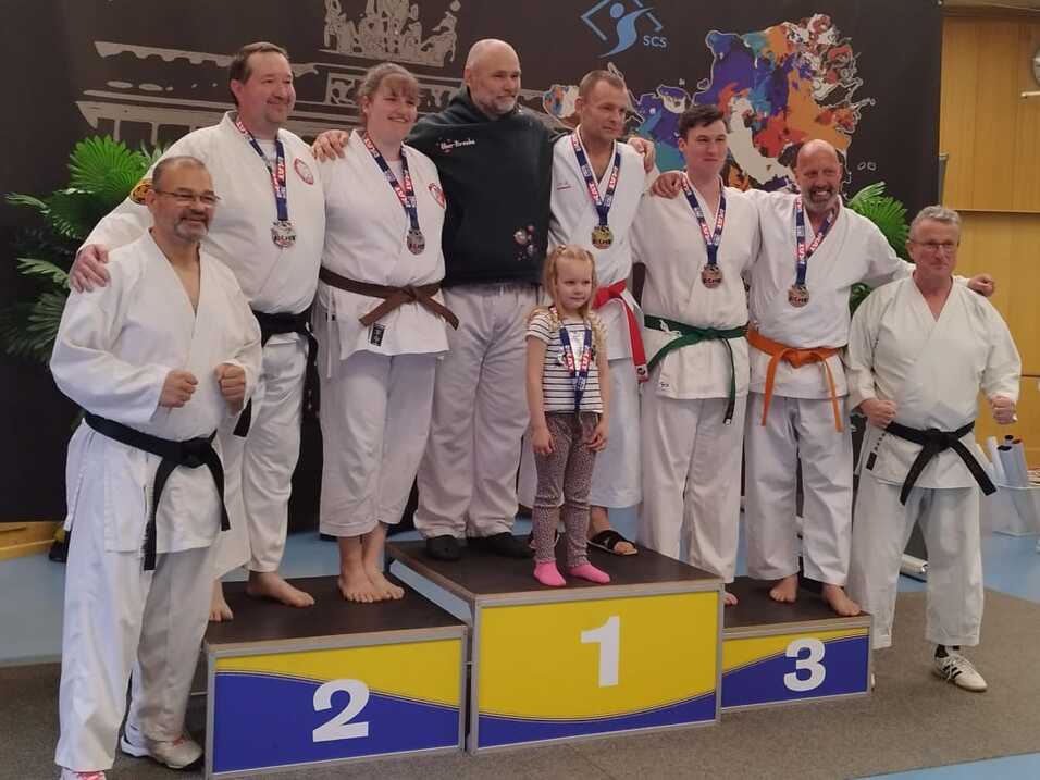 Sieben Karateka in Uniform stehen auf einem Podium mit Medaillen, umgeben von einer dekorativen Kulisse.