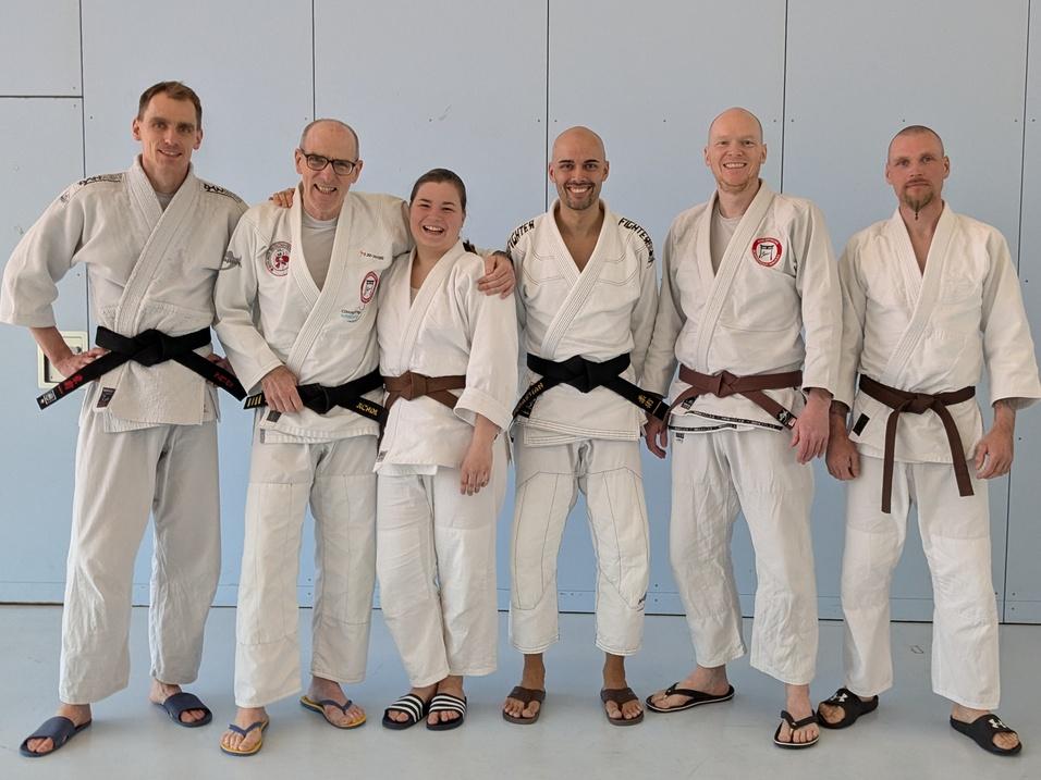 Gruppe von sechs Kampfsportlern in Gi, zwei mit schwarzen, vier mit braunen Gürteln, stehen vor einer blauen Wand.