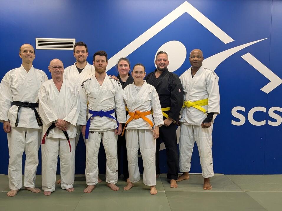 Gruppe von acht Personen in Judokleidung mit verschiedenen Gürtelfarben vor einer blauen Wand des SCS-Studios.