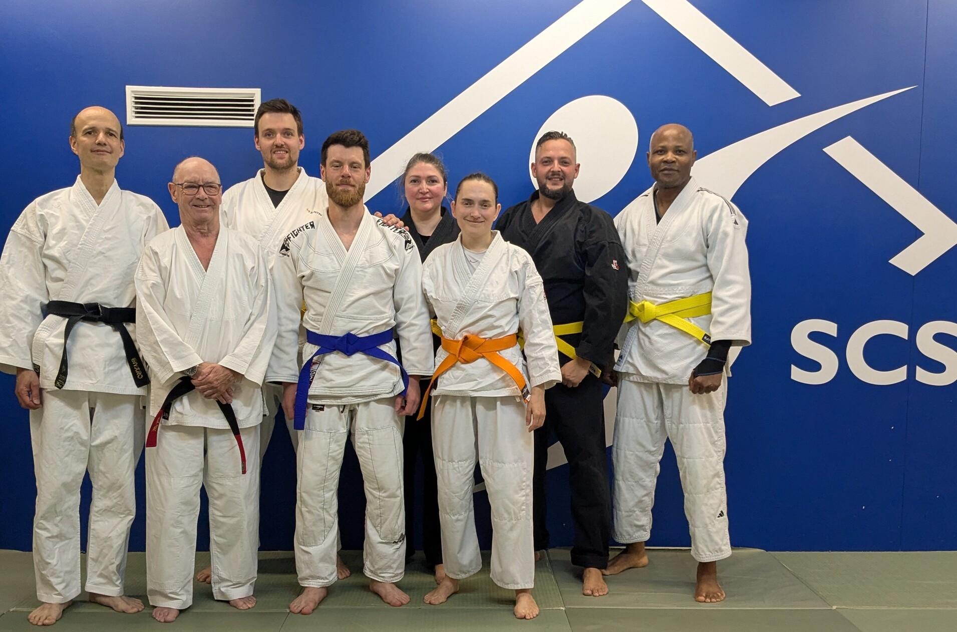 Gruppe von acht Personen in Judokleidung mit verschiedenen Gürtelfarben vor einer blauen Wand des SCS-Studios.