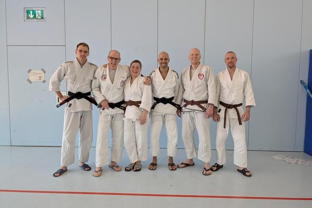 Gruppe von sechs Kampfsportlern in Gi, zwei mit schwarzen, vier mit braunen G&uuml;rteln, stehen vor einer blauen Wand.