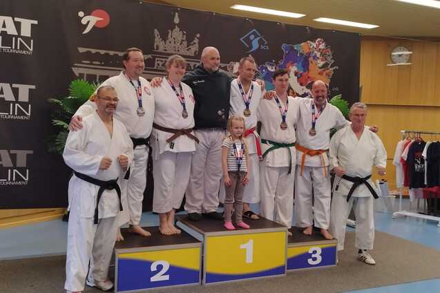 Sieben Karateka in Uniform stehen auf einem Podium mit Medaillen, umgeben von einer dekorativen Kulisse.