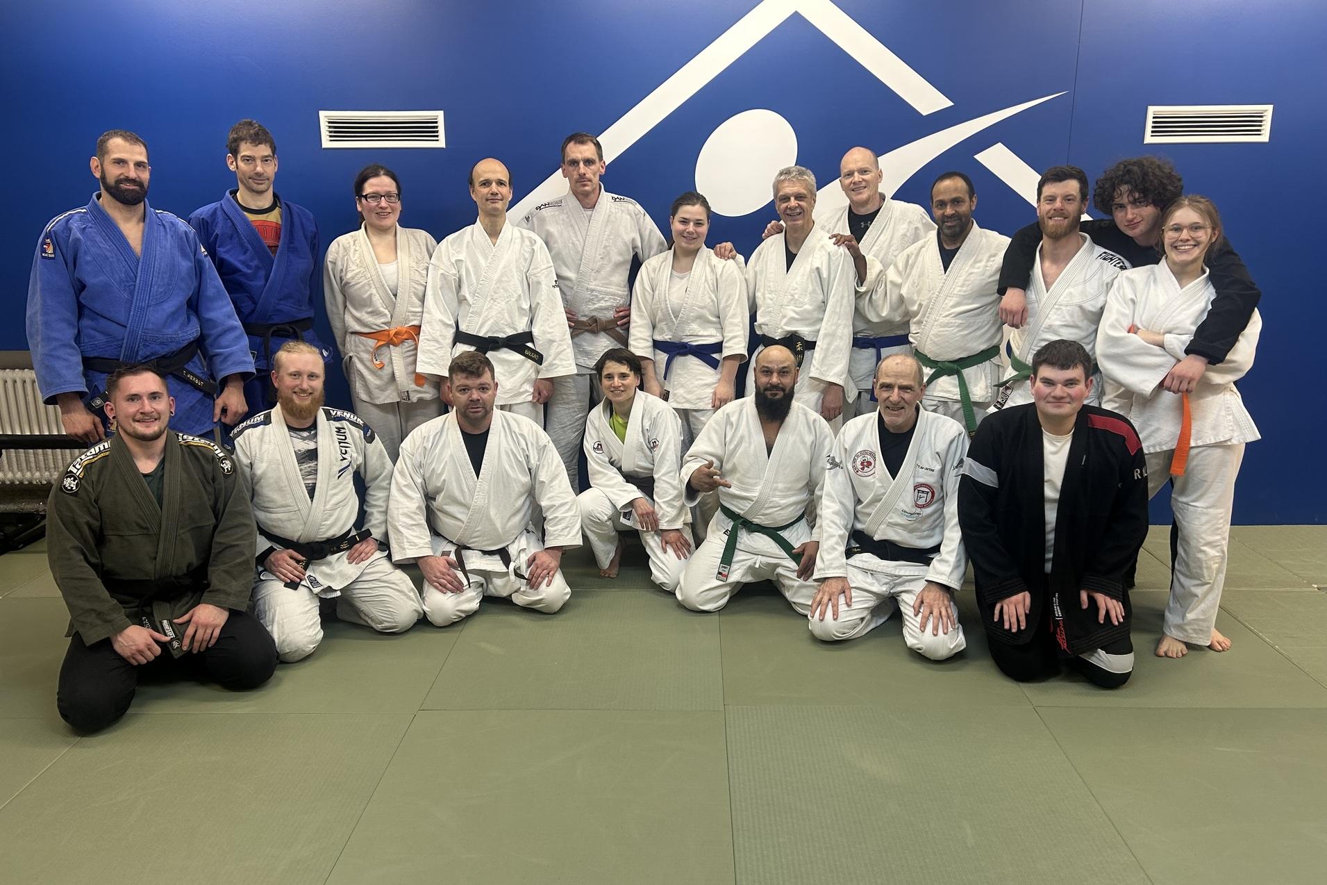 Gruppenfoto von Judo-Praktizierenden in verschiedenen Gürtelstufen auf einer Matte vor einer blauen Wand.