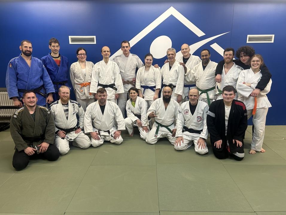 Gruppenfoto von Judo-Praktizierenden in verschiedenen Gürtelstufen auf einer Matte vor einer blauen Wand.