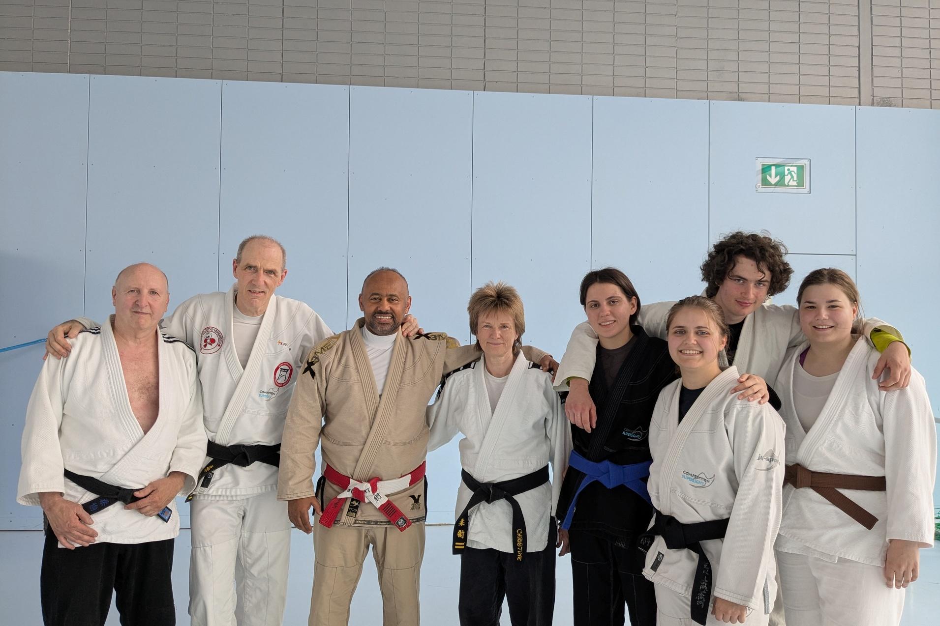 Gruppenfoto von acht Kampfsportlern in Mattenanz&uuml;gen, l&auml;chelnd in einem Trainingsraum mit blauer Wand.