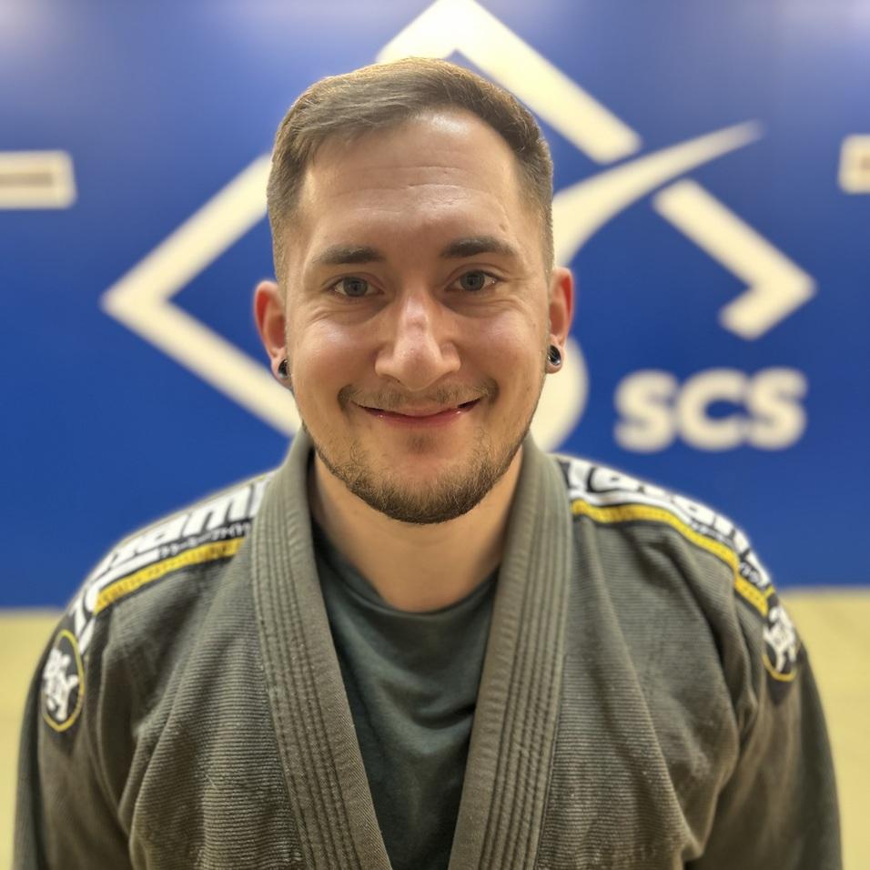 Portait eines lächelnden Mannes in einem grauen BJJ-Gi vor einer blauen Wand mit dem Schriftzug "SCS".