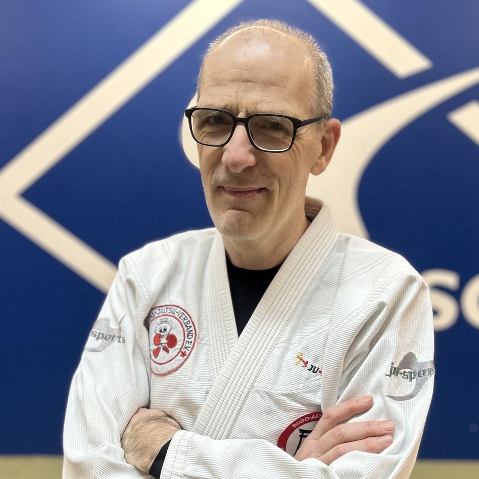 Mann in einem weißen Judoanzug mit schwarzem Gürtel posiert vor einer blauen Wand in einem Trainingsraum.