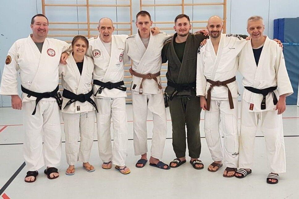 Gruppe von sechs Personen in Kampfsportanzügen, lächelnd, vor einer Sporthalle mit Turngeräten im Hintergrund.