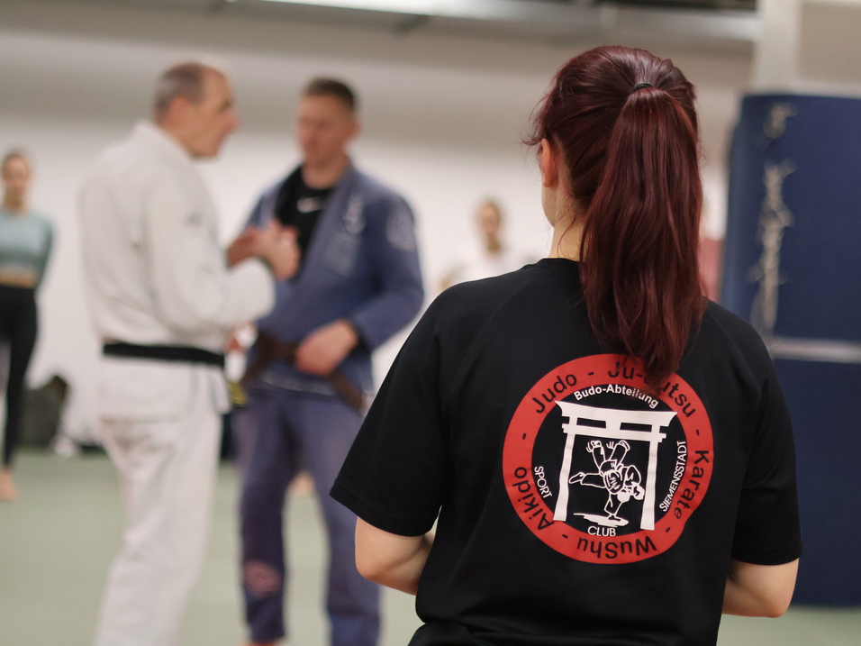 Frau mit langem Haar in T-Shirt mit Kampfsportmotiv beobachtet Judounterricht in einem Trainingsraum.