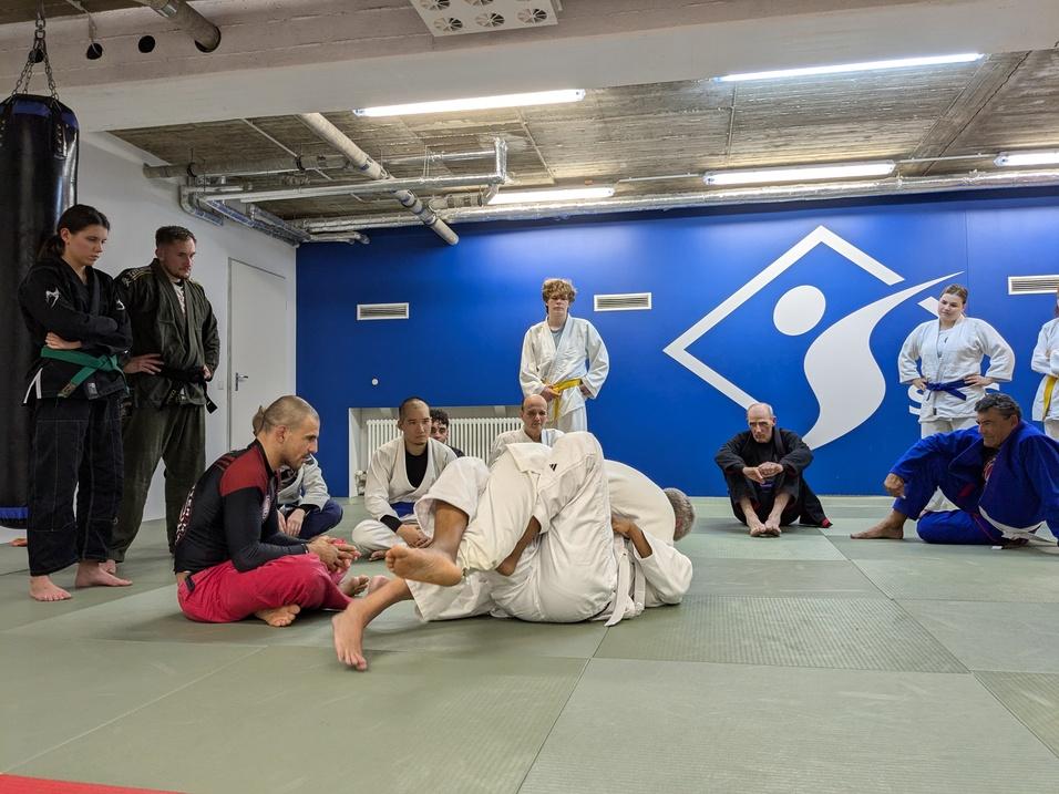 Gruppe von Menschen in Trainingsanzügen beobachtet eine Jiu-Jitsu-Demonstration auf einer Matte in einem Trainingsraum.