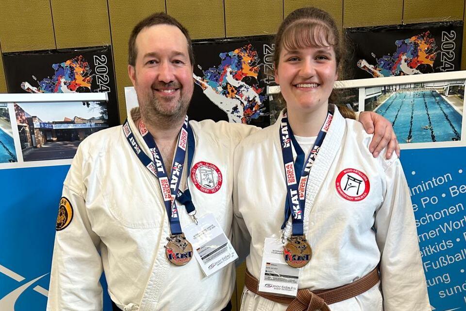 Zwei Karateka mit Medaillen stehen vor einer Wand mit Sportinformationen in einer Sporthalle.