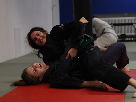 Zwei Frauen im Judo-Anzug üben auf einer Mattenfläche, eine liegt lachend unter der anderen.