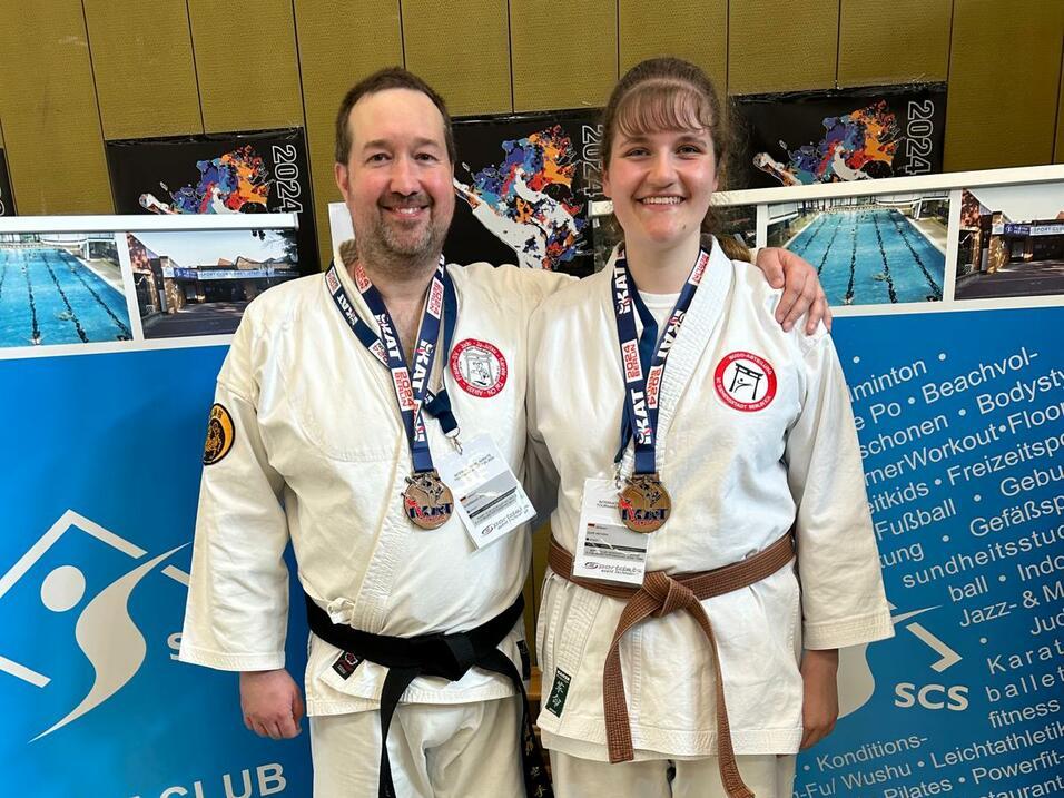 Zwei Karateka mit Medaillen stehen vor einer Wand mit Sportinformationen in einer Sporthalle.