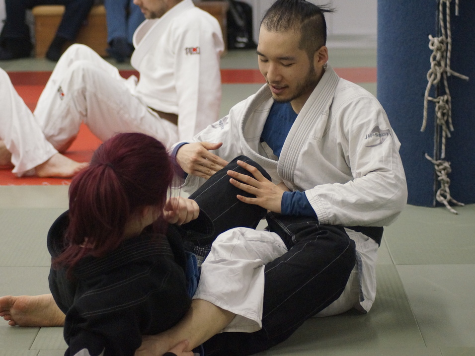 Zwei Personen im Judogi trainieren auf einer Matte, während sie in einer Grappling-Position miteinander interagieren.