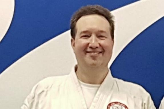 Mann in Karate-Gi hält ein Dan-Diplom und eine Urkunde vor einer blauen Wand.