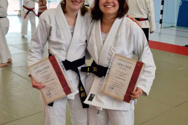 Zwei Judokas in wei&szlig;en Anz&uuml;gen halten stolz Urkunden und l&auml;cheln auf einer Judomatte in einem Sportzentrum.