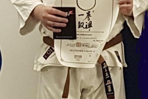 Mann in Karate-Gi h&auml;lt ein Dan-Diplom und eine Urkunde vor einer blauen Wand.