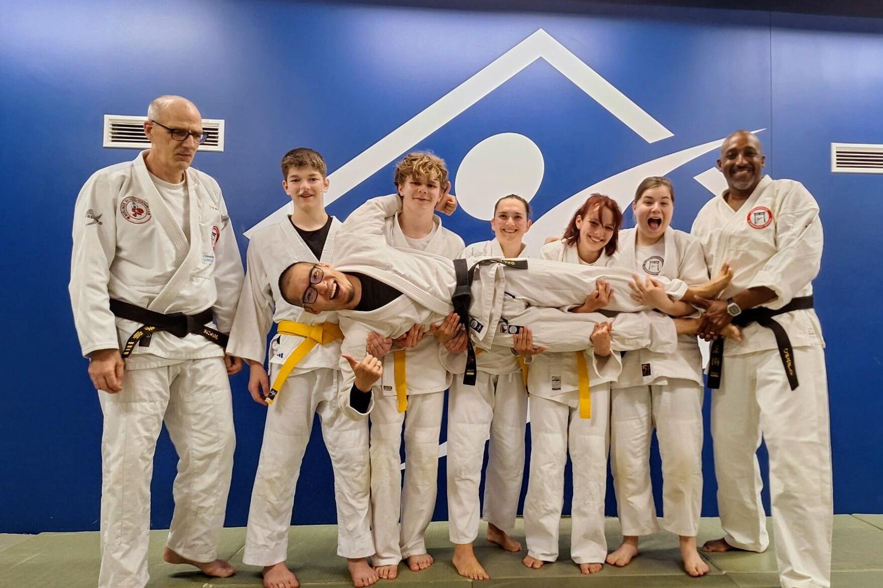 Gruppe von Judokas in wei&szlig;en Anz&uuml;gen, einige lachen, ein Teilnehmer wird von anderen gehalten. Hintergrund in Blau.