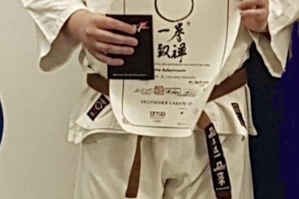 Mann in Karate-Gi h&auml;lt ein Dan-Diplom und eine Urkunde vor einer blauen Wand.