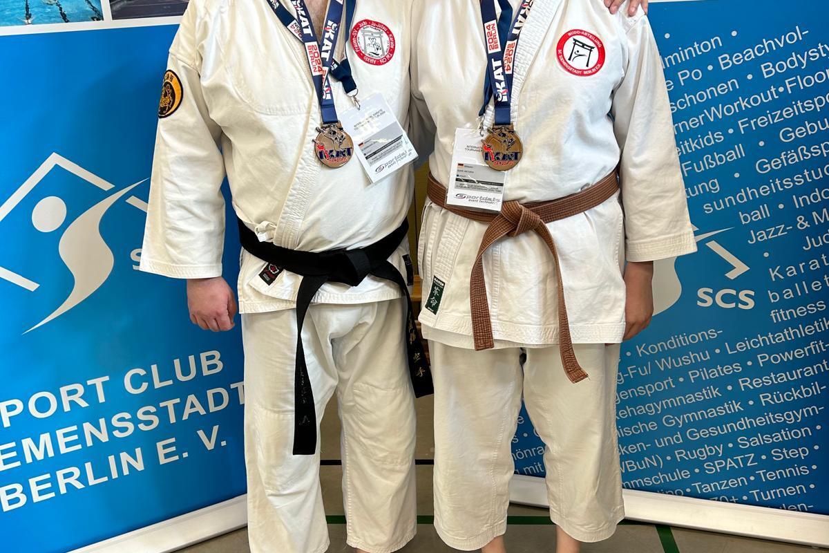 Zwei Karateka mit Medaillen stehen vor einer Wand mit Sportinformationen in einer Sporthalle.