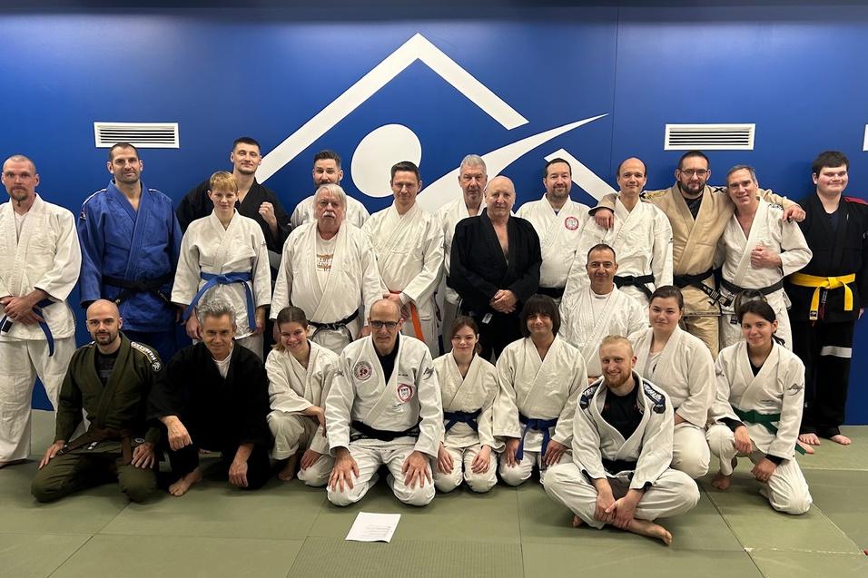 Gruppenfoto von 20 Personen in Judogis auf einer Matte vor blauer Wand mit Logo, einige tragen unterschiedliche Gurtfarben.