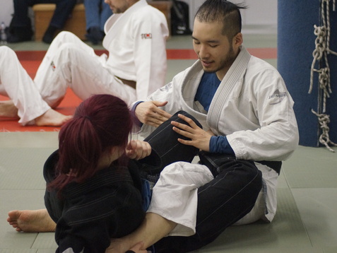 Zwei Personen im Judogi trainieren auf einer Matte, während sie in einer Grappling-Position miteinander interagieren.