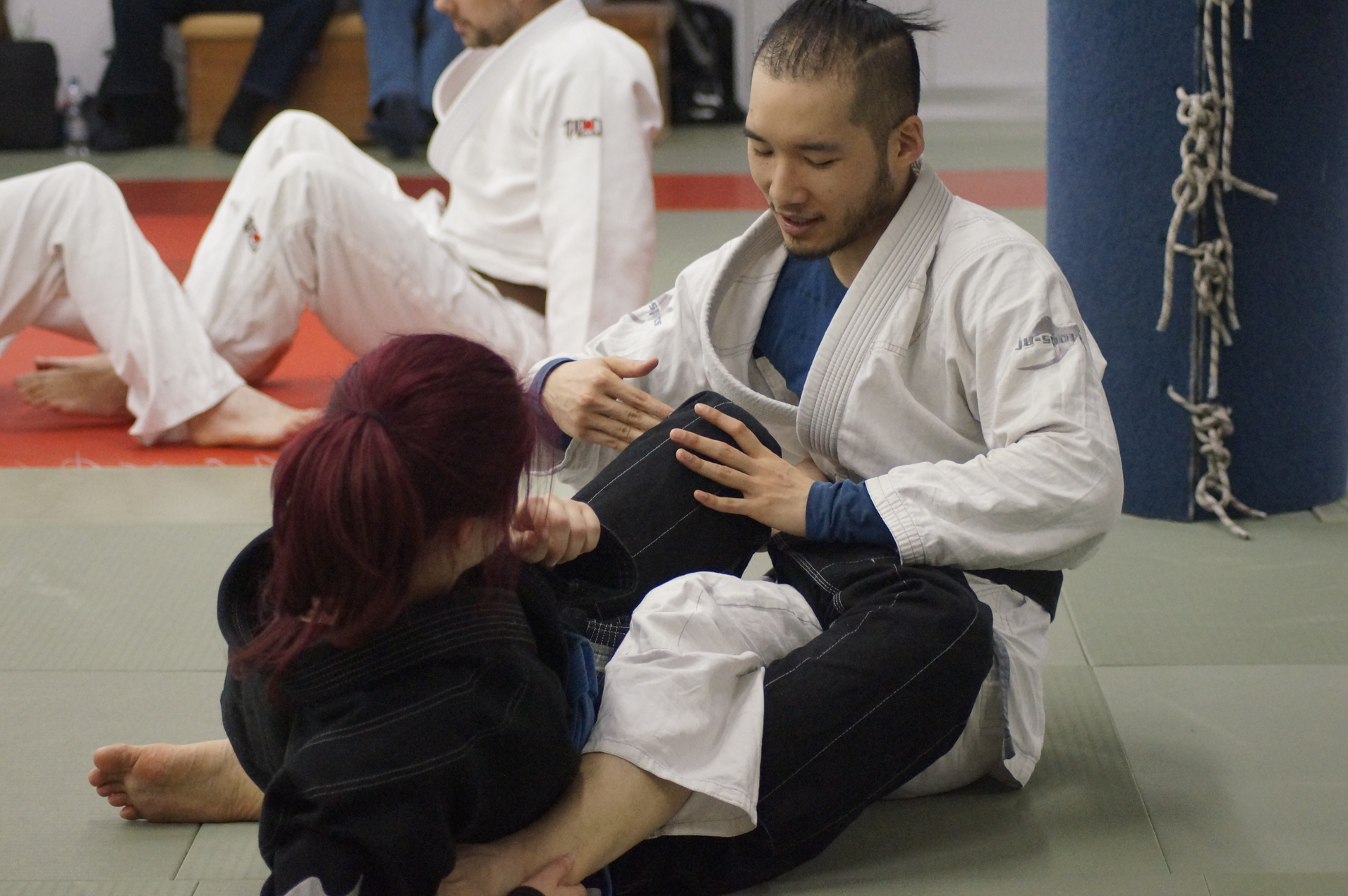 Zwei Personen im Judogi trainieren auf einer Matte, während sie in einer Grappling-Position miteinander interagieren.