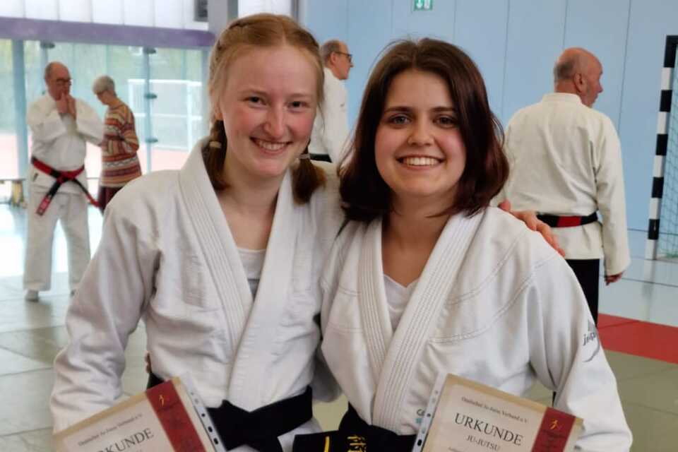 Zwei Judokas in weißen Anzügen halten stolz Urkunden und lächeln auf einer Judomatte in einem Sportzentrum.