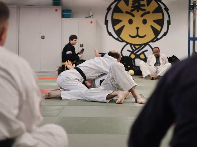 Zwei Kampfsportler in weißen Anzügen trainieren Bodenkampf auf einer Matte, im Hintergrund ein Sportler auf einer Bank.