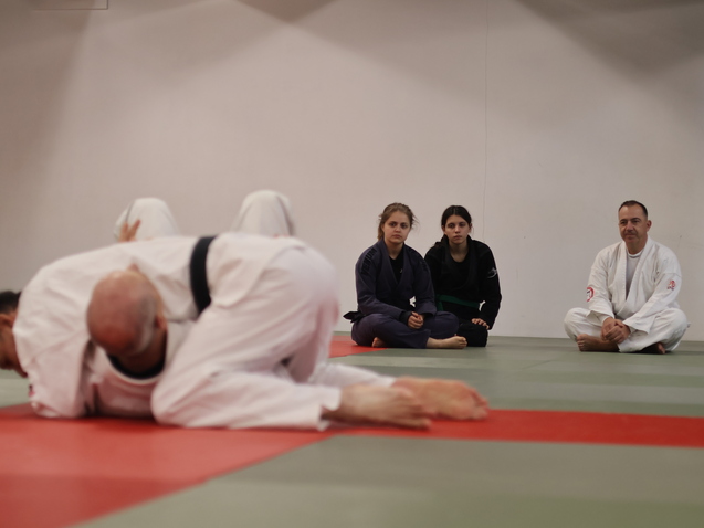 Trainingseinheit im Jiu-Jitsu: Ein Paar übt am Boden, während drei Zuschauer auf einer Matte sitzen und beobachten.