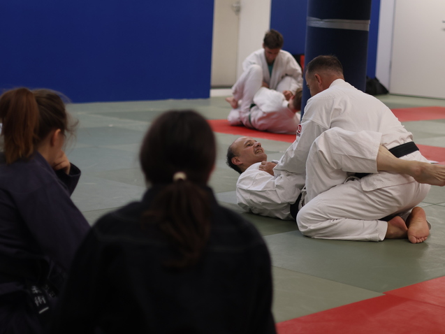 Zwei Judokas im Training, einer auf dem Boden, der andere in einer Kampfposition, mit Zuschauern im Hintergrund.