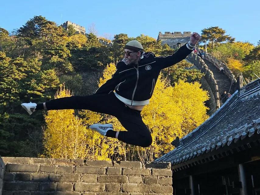 Mann in Sportkleidung springt mit gestrecktem Bein über eine Mauer, im Hintergrund leuchtet die Herbstlandschaft.