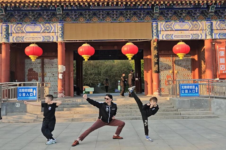 Drei Personen in Karate-Positionen stehen vor einem historischen Geb&auml;ude mit roten Laternen und einem blauem Eingangsschild.