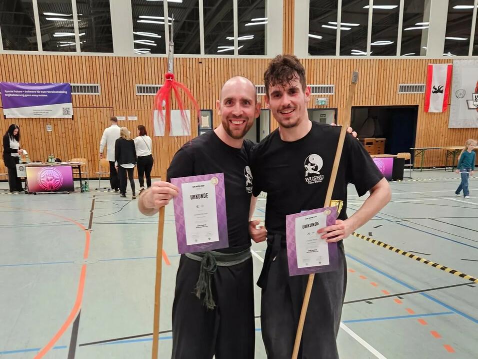 Zwei männliche Sportler in Wushu-Kleidung posieren mit Urkunden und Stäben in einer Sporthalle.