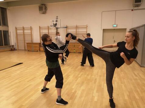 Vier Personen trainieren Kampfsport in einer Sporthalle; eine Jugendlicher führt einen Kick aus.