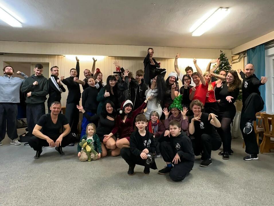 Gruppenfoto von etwa 40 Personen in einem Raum, viele in festlicher Kleidung, lächelnd und posierend um einen Weihnachtsbaum.