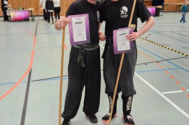 Zwei m&auml;nnliche Sportler in Wushu-Kleidung posieren mit Urkunden und St&auml;ben in einer Sporthalle.