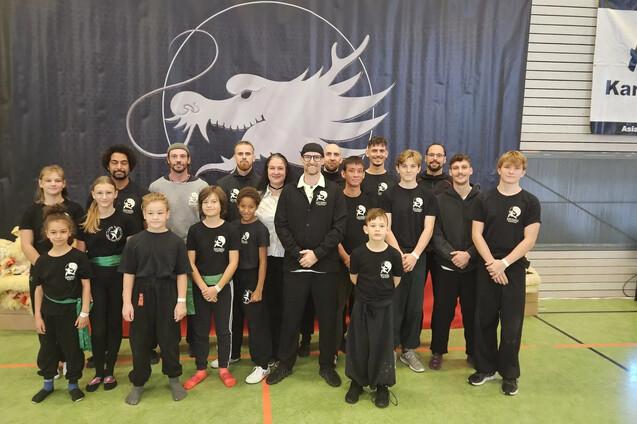Gruppe von Kampfsportlern in Schwarz mit gr&uuml;nem G&uuml;rtel, vor einer Wand mit Drachen-Logo in einer Sporthalle.