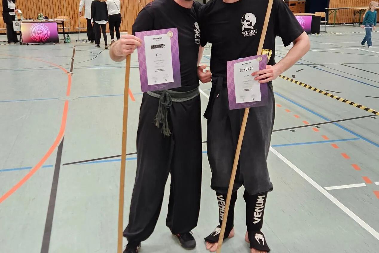 Zwei m&auml;nnliche Sportler in Wushu-Kleidung posieren mit Urkunden und St&auml;ben in einer Sporthalle.