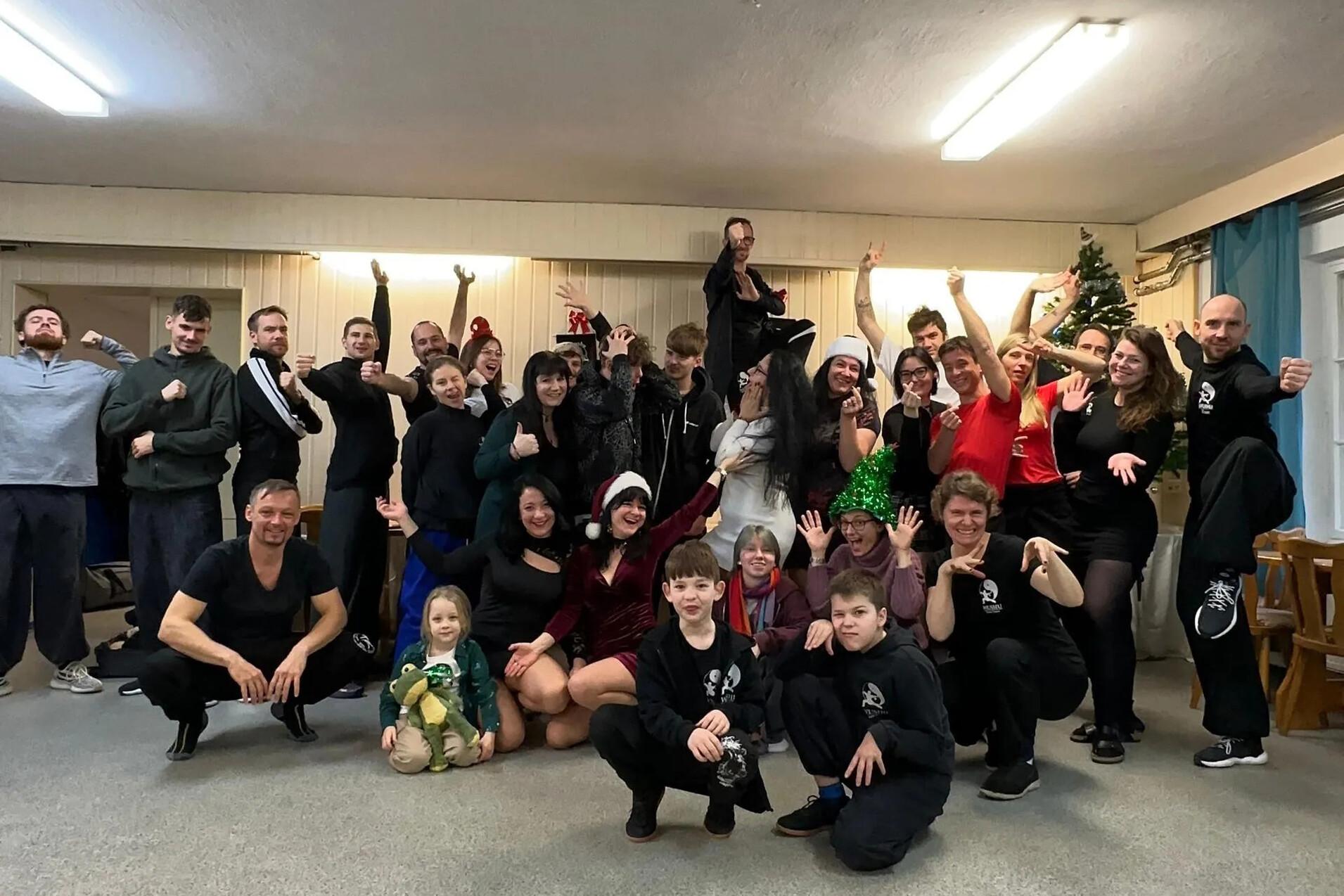 Gruppenfoto von etwa 40 Personen in einem Raum, viele in festlicher Kleidung, l&auml;chelnd und posierend um einen Weihnachtsbaum.