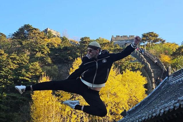 Mann in Sportkleidung springt mit gestrecktem Bein über eine Mauer, im Hintergrund leuchtet die Herbstlandschaft.