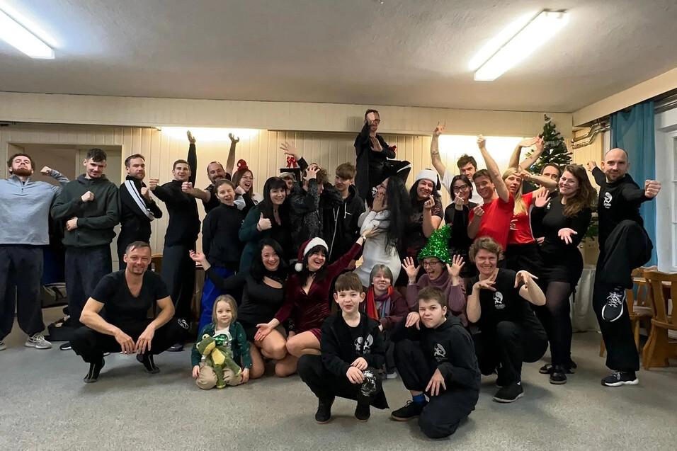 Gruppenfoto von etwa 40 Personen in einem Raum, viele in festlicher Kleidung, l&auml;chelnd und posierend um einen Weihnachtsbaum.