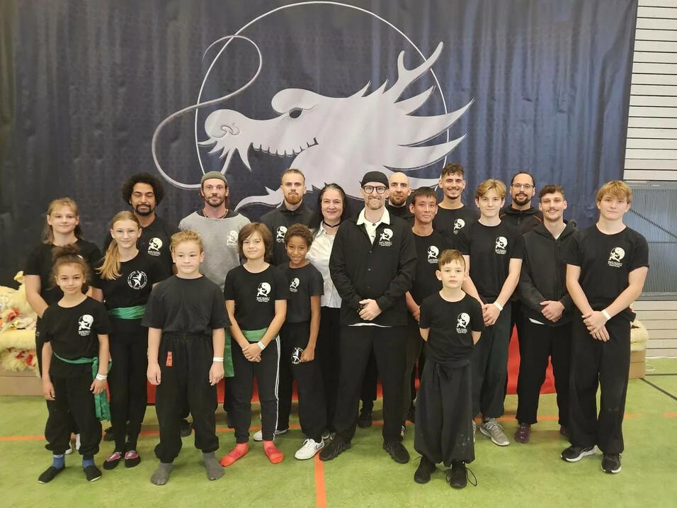 Gruppe von Kampfsportlern in Schwarz mit grünem Gürtel, vor einer Wand mit Drachen-Logo in einer Sporthalle.