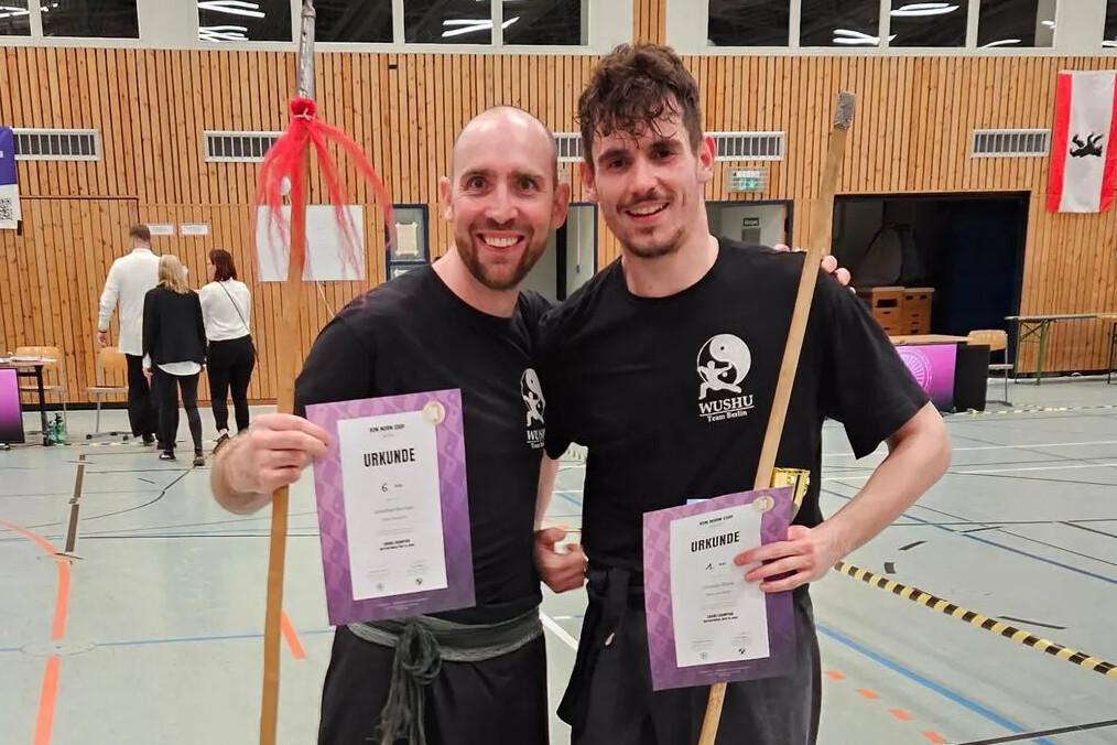 Zwei männliche Sportler in Wushu-Kleidung posieren mit Urkunden und Stäben in einer Sporthalle.