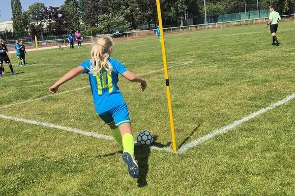 Ein M&auml;dchen in blau-gr&uuml;nem Fu&szlig;balltrikot f&uuml;hrt einen Eckball auf einem Rasenplatz aus, umgeben von Spielern.