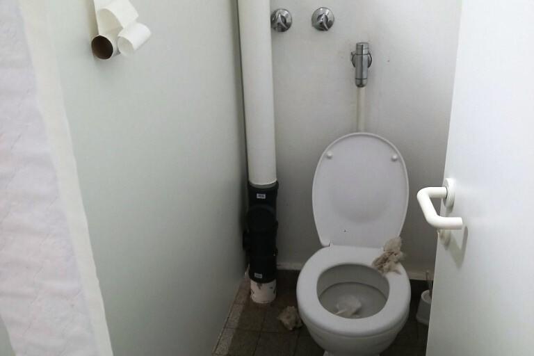 Toilettenraum mit einem Klo, grauen Fliesen und einem Hinweisblatt an der Wand. Sichtbare Abfallstoffe auf dem Boden.