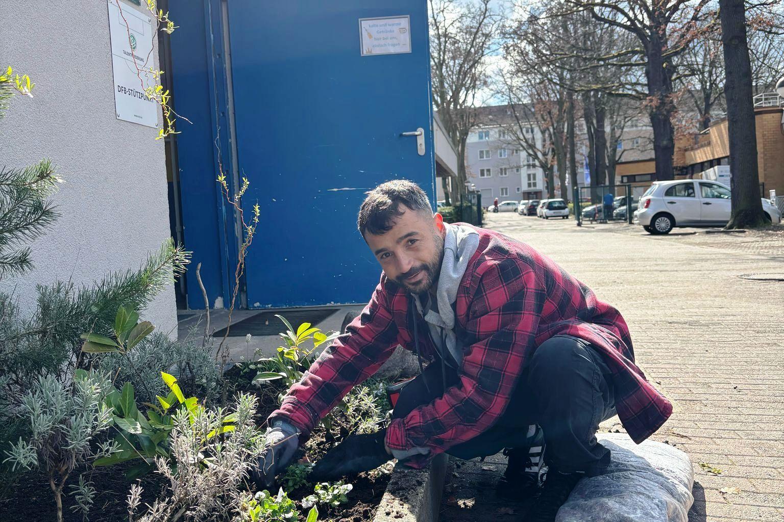 Mann pflanzt Blumen in einem öffentlichen Garten vor einem Gebäude mit blauer Tür unter einem sonnigen Himmel.