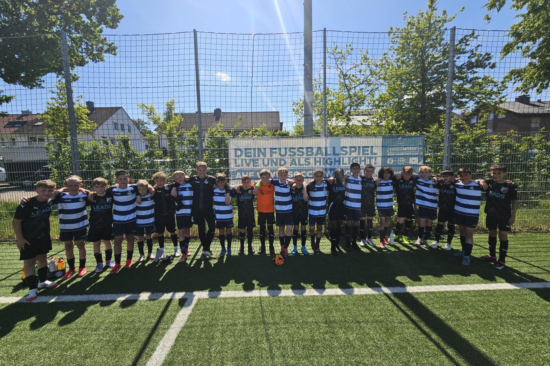 GruppennFoto von Jungen in Fußballtrikots auf einem Kunstrasenplatz, umgeben von einem netz eingezäunten Bereich.