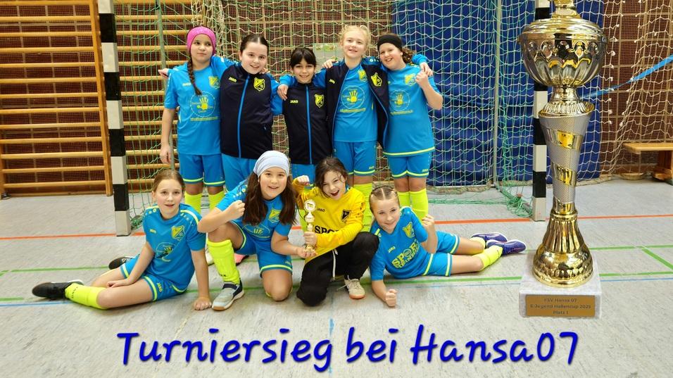 Mädchenfußballmannschaft posiert vor einem Pokal in einer Sporthalle, alle tragen blaue Trikots mit gelben Akzenten.