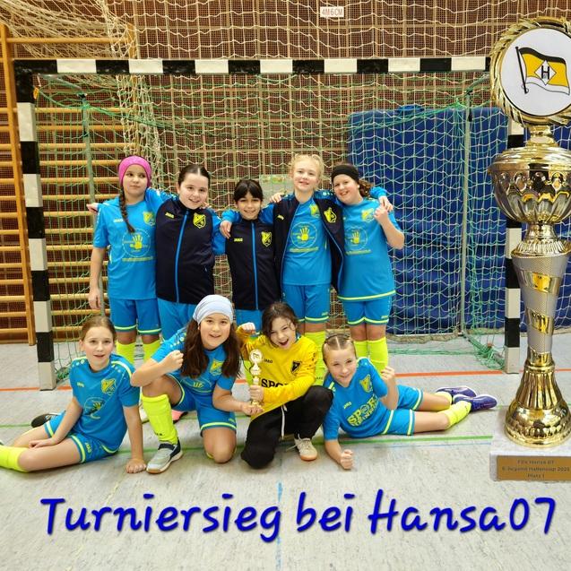 Gruppe junger Fu&szlig;ballspielerinnen posiert mit einem Pokal in einer Sporthalle nach einem Turniersieg.